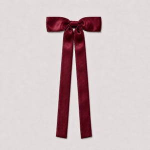May include: A deep burgundy satin bow accessory featuring a central knot and two long, straight, vertical ribbon tails extending downward. The fabric has a smooth, reflective sheen, creating subtle highlights across the surface of the bow.