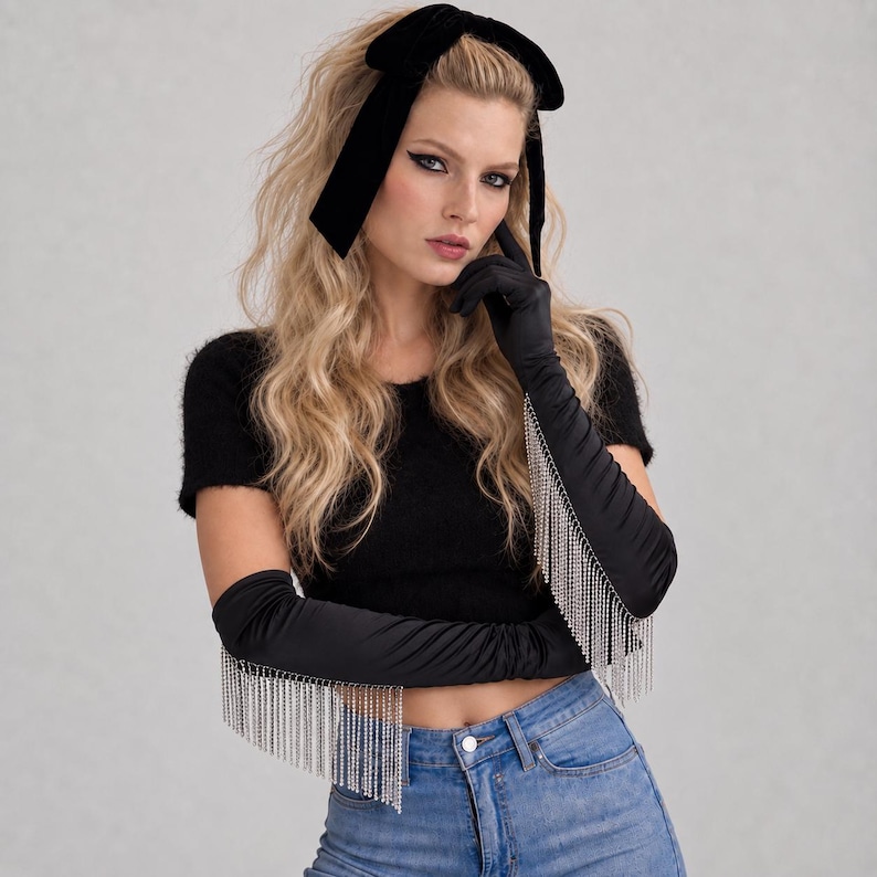 May include: A pair of black satin opera-length gloves featuring long, dangling silver-toned crystal fringe trim along the outer edges. The gloves are paired with a black velvet bow headband, a black short-sleeved fuzzy top, and blue denim jeans.