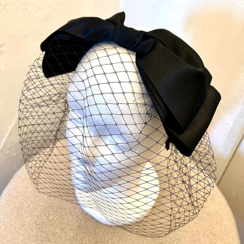 May include: A black satin bow with a black tulle veil. The bow is large and sits on top of the veil. The veil is made of a fine mesh material.