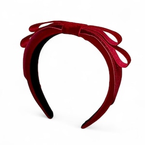 May include: A deep red velvet headband featuring a prominent, structured bow detail at the top. The fabric has a soft, textured appearance with a rich, saturated crimson color throughout the entire band and the bow, creating a uniform look.