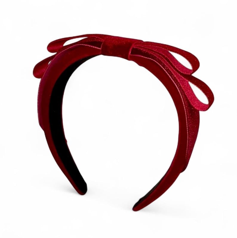 May include: A deep red velvet headband featuring a prominent, structured bow detail at the top. The fabric has a soft, textured appearance with a rich, saturated crimson color throughout the entire band and the bow, creating a uniform look.