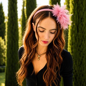 May include: A pink headband featuring a large, fluffy pink feather plume on the side. The headband is worn with a black top and two layered gold necklaces, one with a heart pendant and the other with a circular coin-style charm pendant.