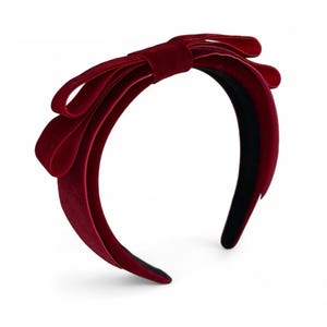 May include: A deep red velvet headband featuring a large, structured bow detail on the side. The headband has a smooth, soft-textured finish in a rich burgundy hue, with a solid black interior lining visible along the inner band structure.