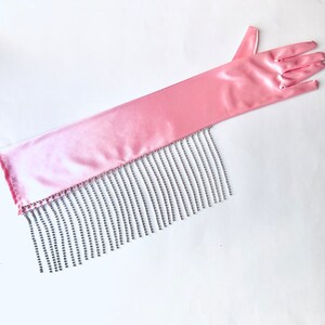 May include: A long, light pink satin opera glove featuring a smooth, lustrous finish. The lower edge of the glove is adorned with a dense fringe of sparkling, clear crystal rhinestones that hang vertically from the fabric hem.