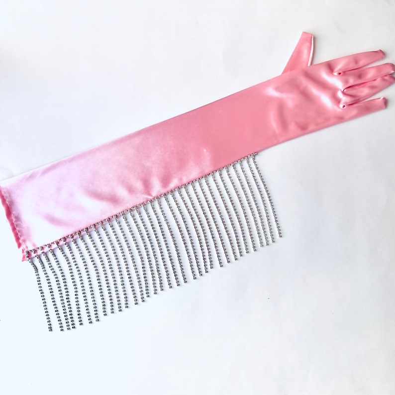 May include: A long, light pink satin opera glove featuring a smooth, lustrous finish. The lower edge of the glove is adorned with a dense fringe of sparkling, clear crystal rhinestones that hang vertically from the fabric hem.