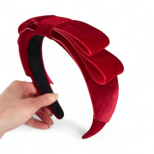 May include: A deep red velvet headband featuring a large, structured bow detail on the side. The headband has a smooth, dark interior lining and a soft, plush texture throughout the exterior surface, creating a bold and uniform crimson appearance.