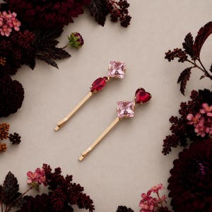 May include: Two gold-tone bobby pins featuring decorative crystal accents. One pin has a square light pink crystal and a marquise-shaped dark red crystal. The other pin features a square light pink crystal and a heart-shaped dark red crystal.