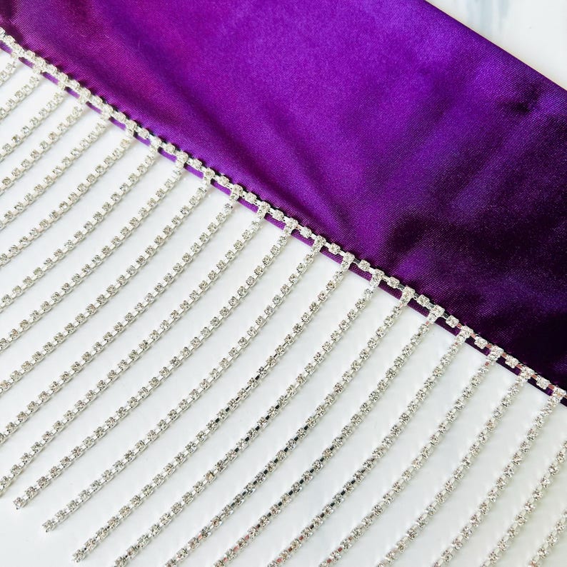 May include: A deep purple satin fabric trim featuring a long, dangling fringe made of multiple strands of clear, sparkling rhinestones set in silver-toned metal. The fringe hangs vertically from the straight edge of the smooth, shiny textile.