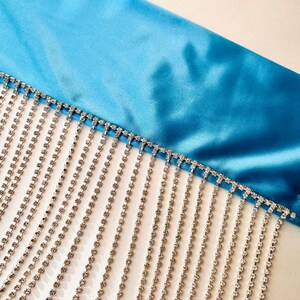 May include: A bright blue satin fabric trim featuring a long, dangling fringe made of multiple vertical strands of clear, faceted rhinestones set in a silver-toned metal chain, creating a shimmering, linear decorative accent.