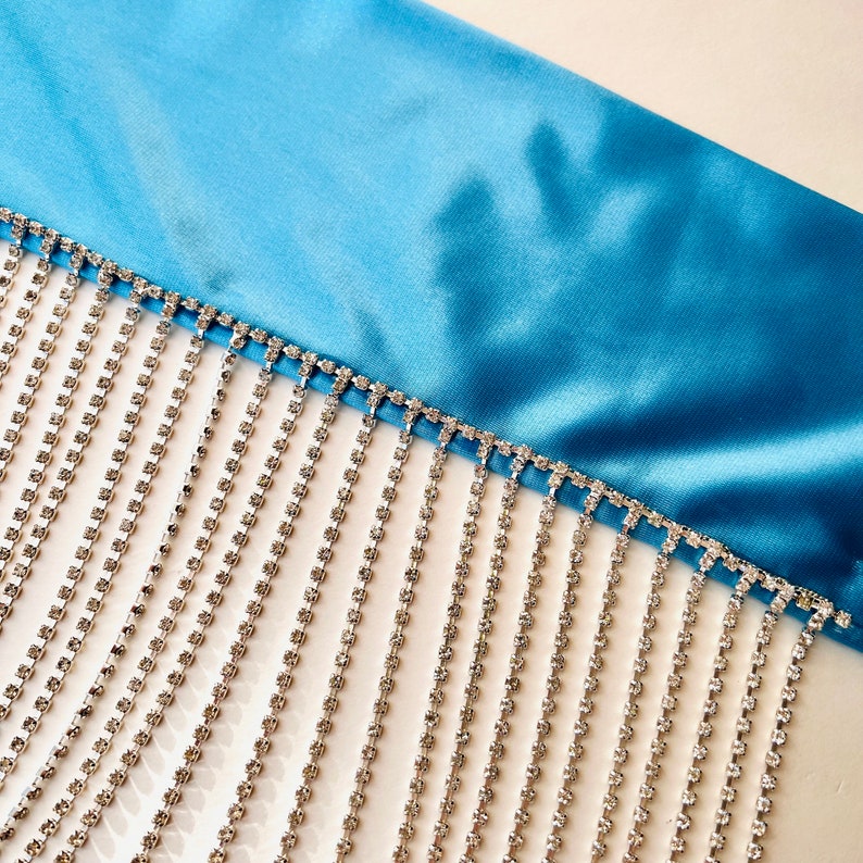 May include: A bright blue satin fabric trim featuring a long, dangling fringe made of multiple vertical strands of clear, faceted rhinestones set in a silver-toned metal chain, creating a shimmering, linear decorative accent.