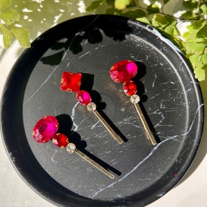 Puede incluir: Cuatro pinzas para el pelo doradas con piedras de imitación rojas y transparentes. Las pinzas están sobre una superficie negra.