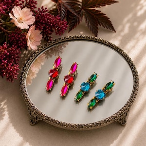 May include: Four hair clips arranged on a round mirror tray. Two clips feature bright pink and red faceted crystals, while the other two display vibrant green and blue faceted crystals. Each clip is set in a gold-toned metal frame.