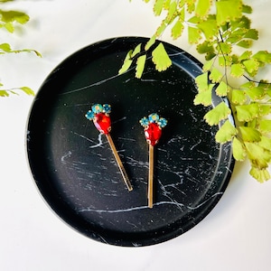 Könnte beinhalten: Zwei goldene Haarklammern mit roten und blauen Kristallverzierungen. Die Klammern befinden sich auf einer schwarzen Marmorfläche.