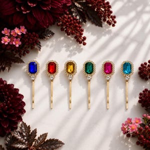 Op de afbeelding: Zes goudkleurige haarspelden op een rij, elk met een rechthoekige gefacetteerde edelsteen in blauw, rood, geel, groen, roze of lichtblauw. Elke steen is omringd door kleine heldere kristallen en een enkel kristallen accent.