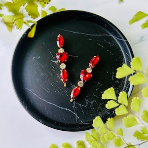 May include: Two gold hair clips with red and clear rhinestones. The clips are on a black marble surface.