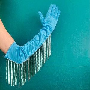 May include: A light blue satin opera-length glove featuring a long fringe of clear, sparkling rhinestones attached along the outer edge. The smooth, shiny fabric extends past the elbow, while the dangling crystal strands create a shimmering effect.