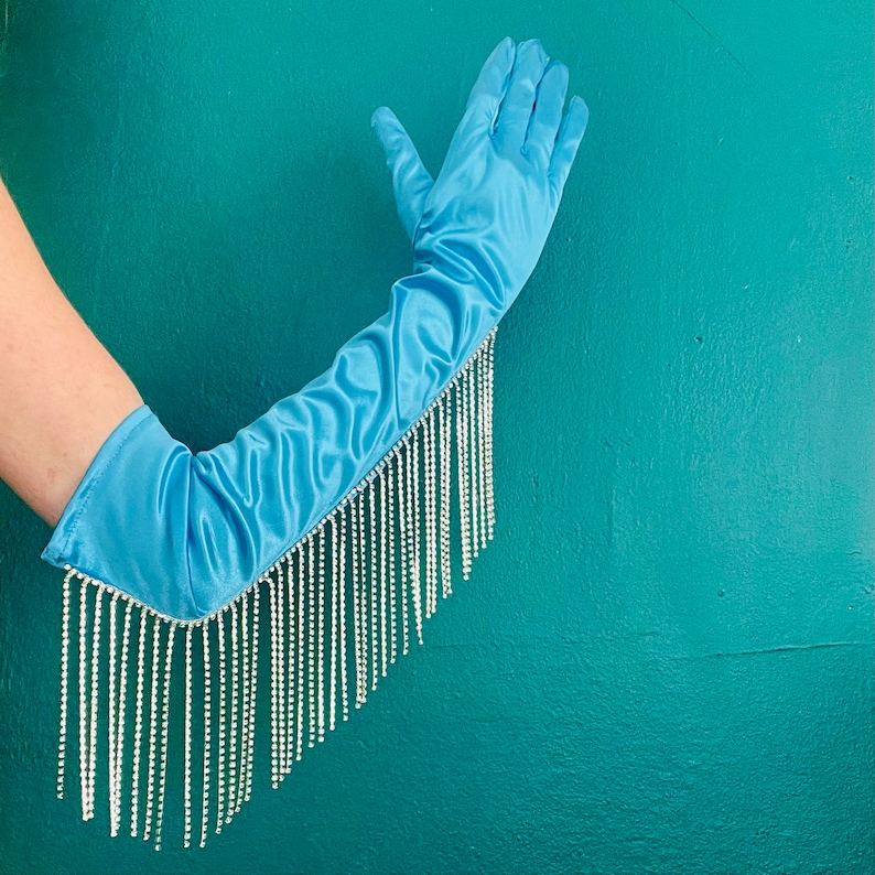 May include: A light blue satin opera-length glove featuring a long fringe of clear, sparkling rhinestones attached along the outer edge. The smooth, shiny fabric extends past the elbow, while the dangling crystal strands create a shimmering effect.