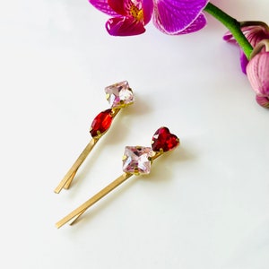 May include: Two gold hair clips with pink and red crystal embellishments. One clip has a square pink crystal and a red teardrop crystal. The other clip has a square pink crystal and a red heart-shaped crystal.
