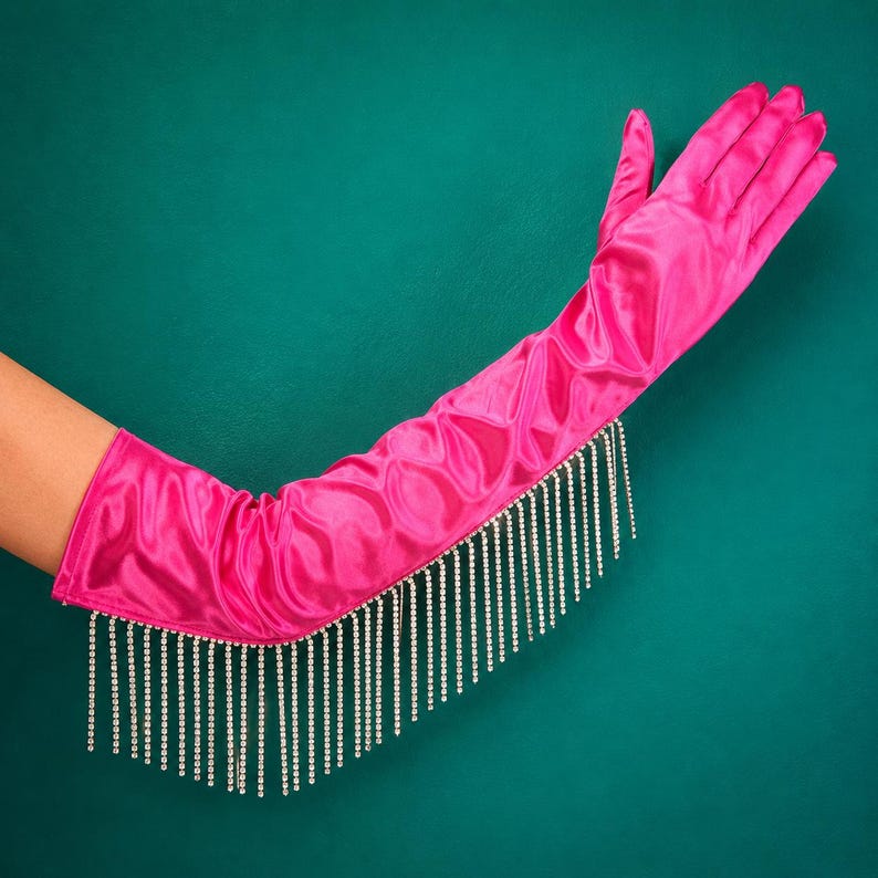 May include: An opera-length hot pink satin glove featuring a long fringe of clear, sparkling rhinestones attached along the outer edge. The smooth, shiny fabric extends from the fingers up to the forearm, accented by the dangling crystal trim.
