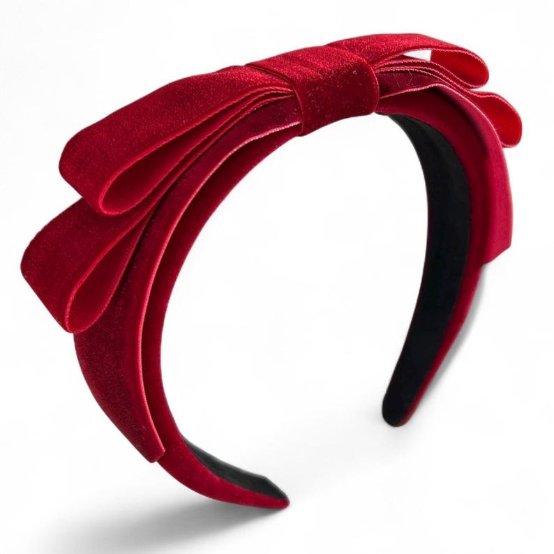 May include: A deep red velvet headband featuring a large, multi-layered bow detail on the top. The headband has a smooth, rounded band structure with a dark interior lining, showcasing a rich, soft-textured fabric finish throughout the design.