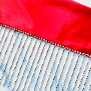 May include: A vibrant red fabric border featuring a long, dangling fringe made of multiple vertical strands of clear, sparkling rhinestones. The silver-toned metal settings hold each individual stone in a uniform, linear arrangement.
