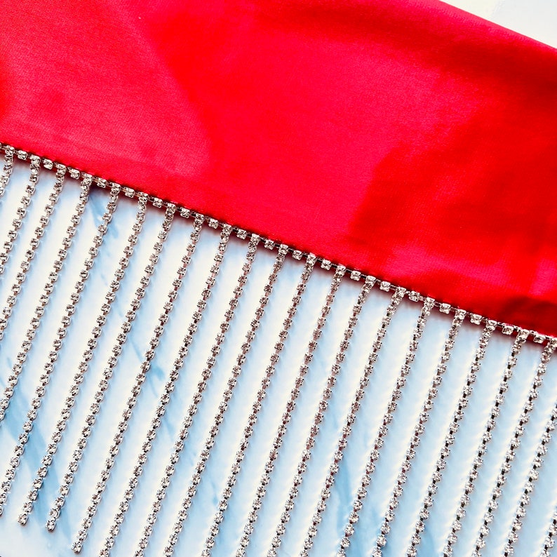 May include: A vibrant red fabric border featuring a long, dangling fringe made of multiple vertical strands of clear, sparkling rhinestones. The silver-toned metal settings hold each individual stone in a uniform, linear arrangement.