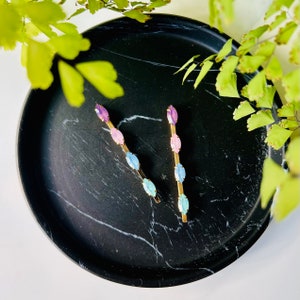 Puede incluir: Dos pinzas para el pelo doradas con detalles de piedras preciosas azul pastel, rosa y morado sobre una superficie de mármol negro.