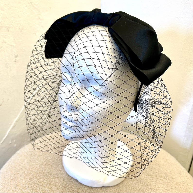 May include: A black satin bow headband with a black tulle veil attached. The headband is worn on a white mannequin head.