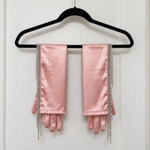 May include: A pair of elbow-length satin gloves in a soft pink hue. Each glove features a long, vertical fringe of clear, sparkling rhinestones attached along the outer edge, extending from the wrist down past the fingertips.