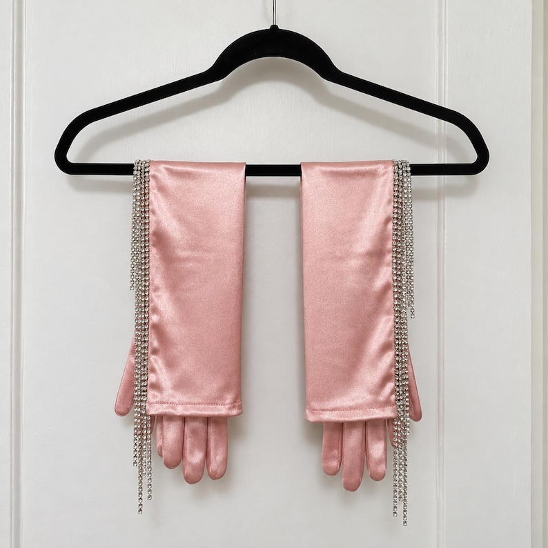 May include: A pair of elbow-length satin gloves in a soft pink hue. Each glove features a long, vertical fringe of clear, sparkling rhinestones attached along the outer edge, extending from the wrist down past the fingertips.