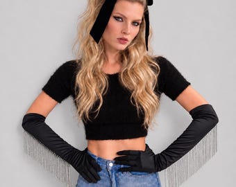 May include: Black opera-length satin gloves featuring long silver-toned crystal fringe trim along the outer forearms. The gloves are paired with a black fuzzy short-sleeve crop top and a black velvet hair bow headband.