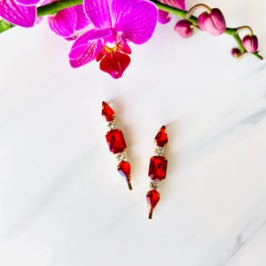 May include: A pair of red and gold earrings with a geometric design. The earrings feature three red stones, two teardrop shaped and one rectangular, with clear rhinestones between each stone.