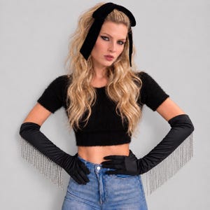 May include: Black opera-length satin gloves featuring long silver-toned crystal fringe trim along the outer forearms. The gloves are paired with a black fuzzy short-sleeve crop top and a black velvet hair bow headband.