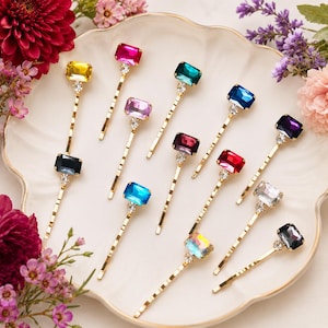 Può includere: Un set di dieci forcine dorate, ognuna caratterizzata da una pietra preziosa rettangolare sfaccettata in vari colori tra cui giallo, rosa, verde acqua, blu, viola, rosso e trasparente, arricchita da un piccolo cristallo chiaro alla base.