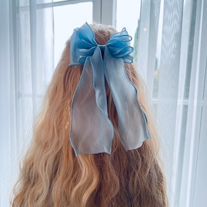 Puede incluir: Un lazo para el pelo de organza azul claro con largas cintas sueltas. El lazo está atado al cabello. La tela transparente tiene una apariencia delicada y aireada, perfecta para añadir un toque de elegancia a cualquier peinado.