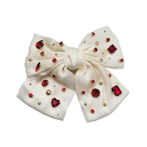 May include: A white satin bow with red and gold embellishments. The bow is decorated with various shapes and sizes of red rhinestones and gold beads.