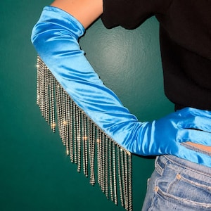 May include: A pair of bright blue satin opera-length gloves featuring long, dangling silver-toned rhinestone fringe that extends from the forearm down to the wrist, creating a shimmering effect against the smooth, reflective fabric of the gloves.