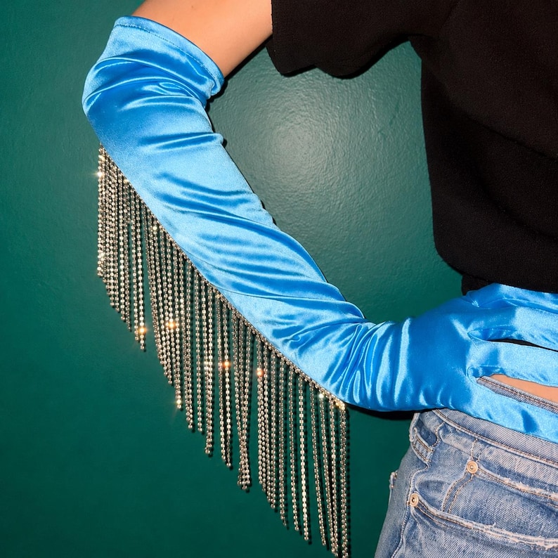 May include: A bright blue satin opera-length glove featuring a long, cascading fringe of clear, reflective rhinestones attached along the outer edge of the forearm, extending down from the elbow toward the wrist area.
