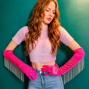 May include: A pair of vibrant hot pink satin opera-length gloves featuring long, dangling silver rhinestone fringe trim along the outer edges. The gloves extend past the elbows and are made of a smooth, shiny fabric that catches the light.