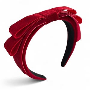 May include: A deep red velvet headband featuring a large, multi-layered bow detail on the top. The headband has a smooth, dark interior lining and a soft, plush texture throughout the exterior surface, creating a consistent monochromatic appearance.