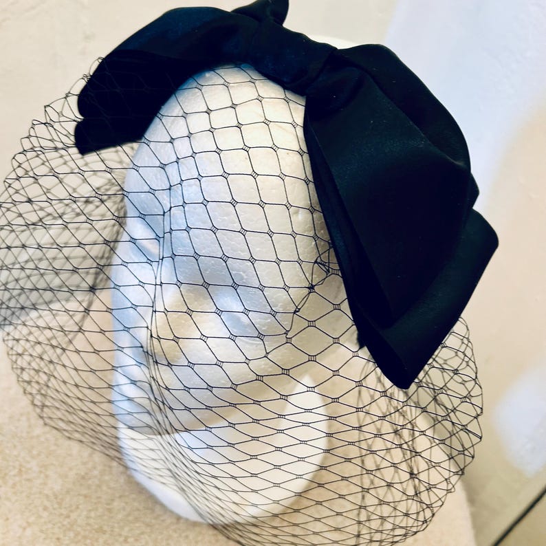 May include: A black satin bow with a black birdcage veil. The veil is made of a fine mesh material.