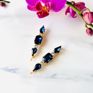 May include: Two gold hair clips with blue and clear rhinestones. The clips are arranged on a white surface.