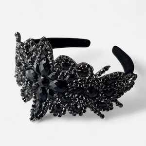 May include: A black headband featuring an ornate, asymmetrical arrangement of faceted black crystals and beads. The decorative section is densely covered in various sizes of dark, reflective stones, creating a textured, layered appearance.