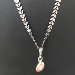 Könnte beinhalten: Eine silberne Halskette mit einer blattförmigen Kette und einem kleinen, runden, rosafarbenen Anhänger.