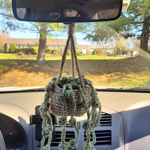 May include: A crocheted hanging plant with green and brown yarn, hanging from the rearview mirror of a car. The plant has a small pot and long strands of yarn that resemble vines.