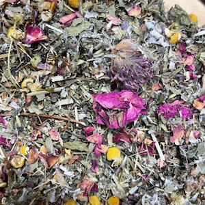 May include: A close-up of a mixture of dried herbs and flowers, including lavender, chamomile, rose petals, and clover. The mixture is a light green color with pops of purple, pink, and yellow.