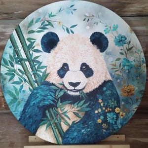 Könnte beinhalten: Ein runder Wandteller mit einem detaillierten Gemälde eines Pandas, der grüne Bambusstangen hält. Der Panda ist von blauen und gelben Blumen vor einem weichen, hellen Hintergrund umgeben. Das Kunstwerk steht auf einem Holzständer.