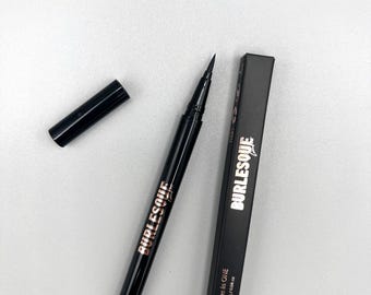 May include: A black eyeliner pen with a matching cap and a rectangular box. The pen and box both feature the word BURLESQUE in white lettering. The box displays the text Eyeliner Glue in ONE and the net weight of 1.2g/0.04 oz.