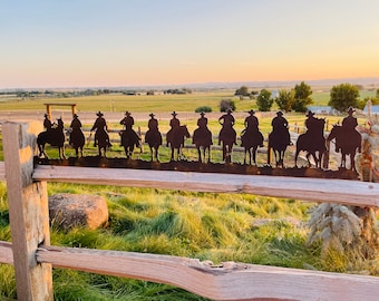 Könnte beinhalten: Eine Metallsilhouette einer Reihe von Cowboys, die auf Pferden reiten, montiert auf einem Holzzaun vor dem Hintergrund eines grasbewachsenen Feldes und eines fernen Horizonts.