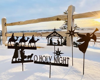May include: A set of six metal Christmas yard decorations, including a nativity scene, three wise men riding camels, a kneeling angel, and a sign that reads "O Holy Night". The decorations are silhouetted against a snowy background.